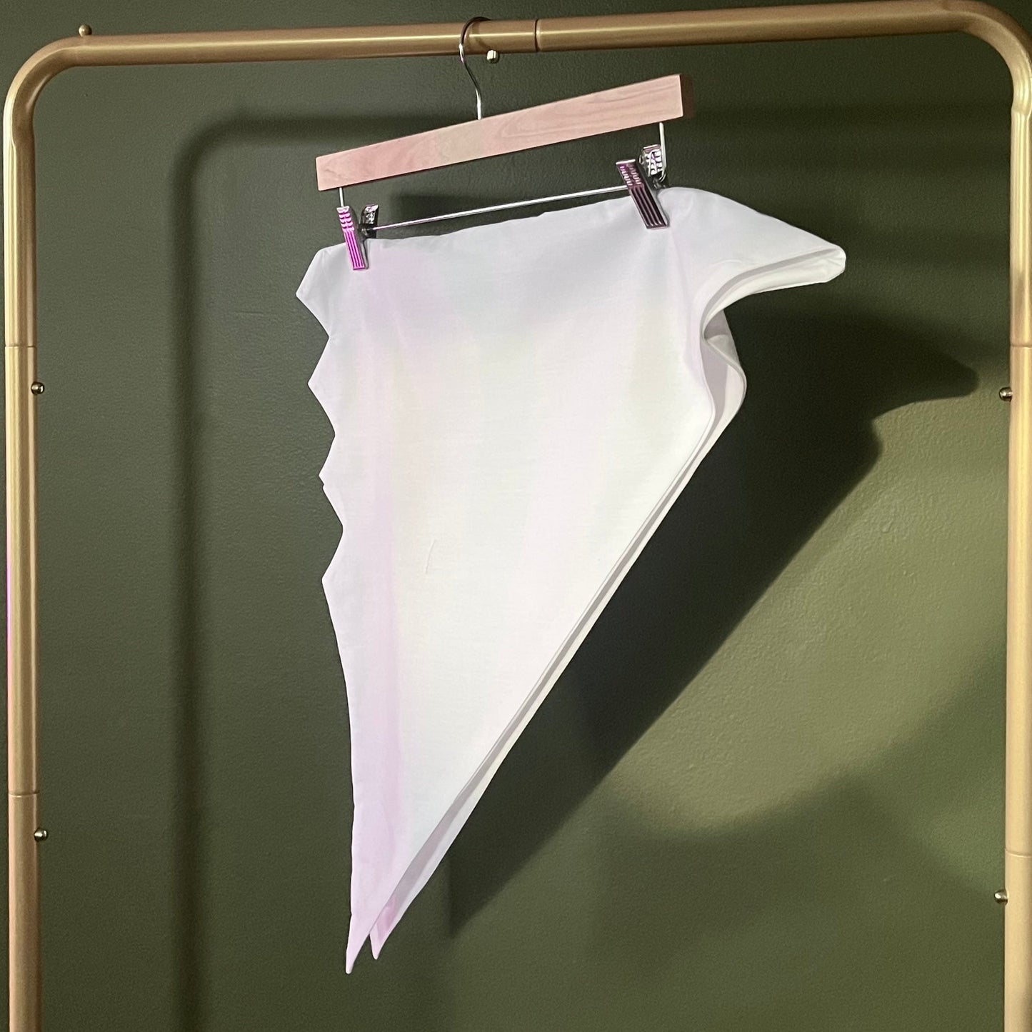 A white triangular head tie with a zig zag edge, hanging from a wooden hanger on a gold-colored clothing rack against a dark olive green wall.